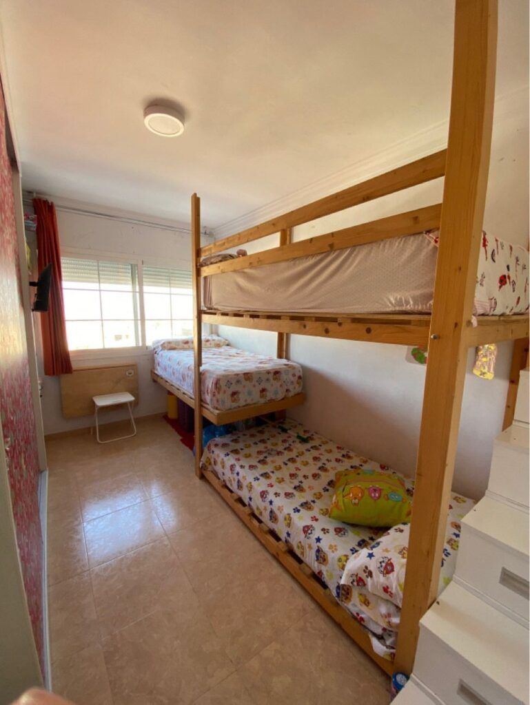 Un dormitorio luminoso con suelo de baldosas, que presenta una litera de madera con ropa de cama de colores, una cama individual junto a la ventana, un pequeño taburete blanco, cortinas rojas y escaleras que conducen a la litera superior. La luz del sol entra a raudales por la ventana.