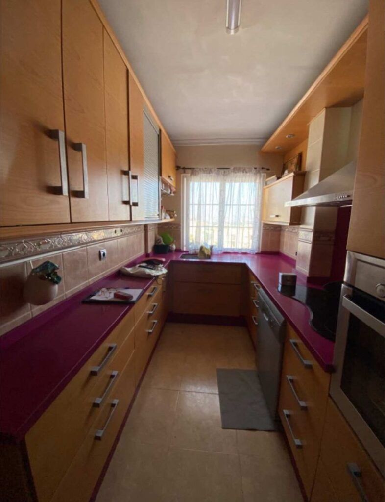 Una cocina estrecha con armarios de madera, encimeras rosas, electrodomésticos de acero inoxidable, un lavavajillas y una ventana en el extremo que deja pasar la luz natural. En el suelo de baldosas, cerca del fregadero, hay una pequeña alfombrilla.
