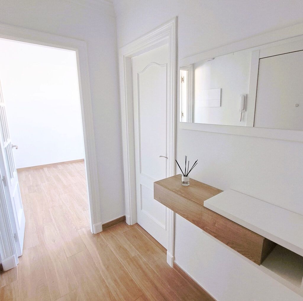 Un pasillo luminoso con suelo de madera clara, paredes blancas, dos puertas blancas cerradas, un portal abierto y una estantería flotante de madera que sostiene un difusor de carrizo bajo un gran espejo.