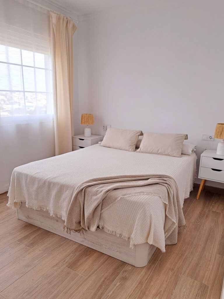 Un dormitorio luminoso y minimalista con una cama doble cubierta con ropa de cama beige, dos almohadas y una manta. Hay mesillas de noche a juego con lámparas a ambos lados, suelos de madera y cortinas transparentes sobre una gran ventana.