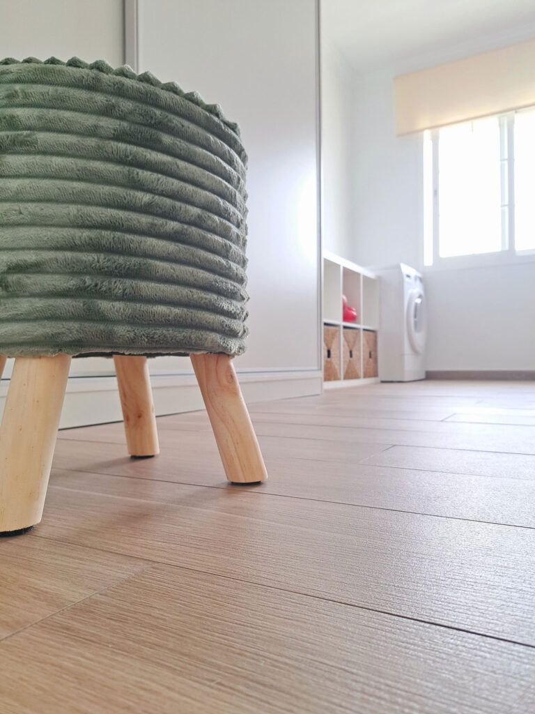 Primer plano de un taburete verde con textura y patas de madera sobre un suelo de madera clara en un luminoso y moderno lavadero con armarios blancos y una lavadora cerca de una ventana.