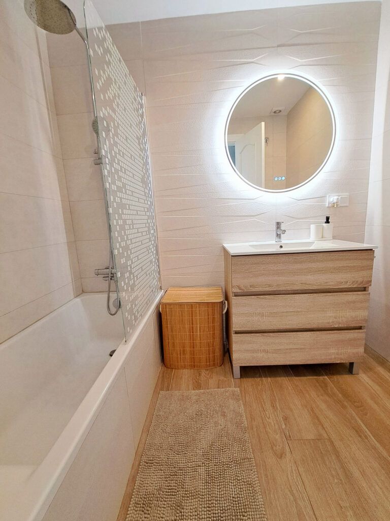 Un cuarto de baño moderno con suelo de madera clara, una bañera con mampara de cristal punteado, un tocador de madera con lavabo, un espejo redondo retroiluminado y un cesto de ropa sucia tejido junto al tocador.