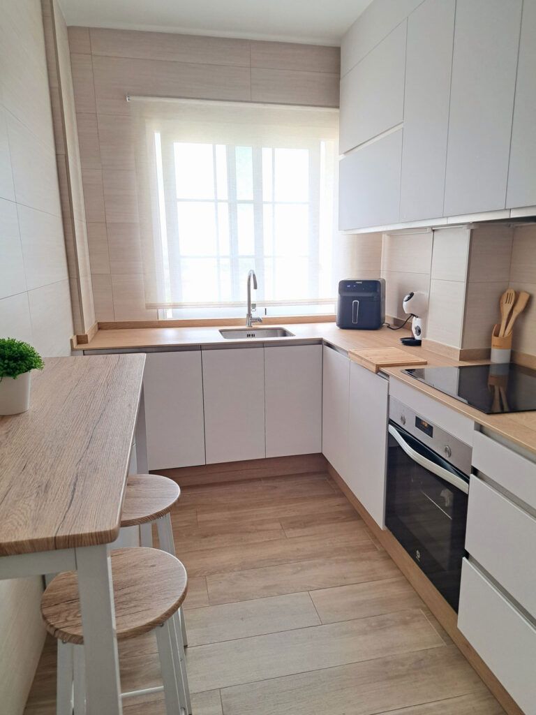 Cocina moderna y minimalista con armarios blancos, encimeras de madera, un horno empotrado, un fregadero bajo una ventana luminosa, dos taburetes en una mesa estrecha y pequeños electrodomésticos sobre la encimera. El suelo y la decoración de madera clara añaden calidez.