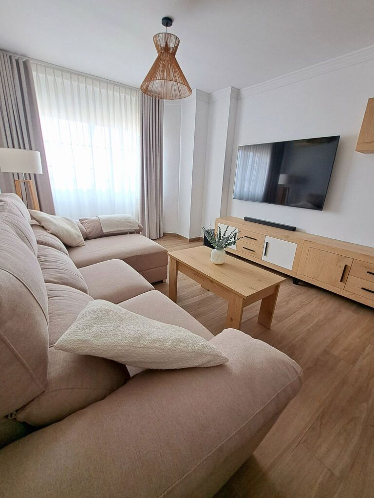 Un moderno salón con un sofá seccional beige claro, una mesa de centro de madera, una maceta, un gran televisor de pantalla plana colgado en la pared, un soporte de madera para el televisor y cortinas transparentes en una ventana por la que entra luz natural.