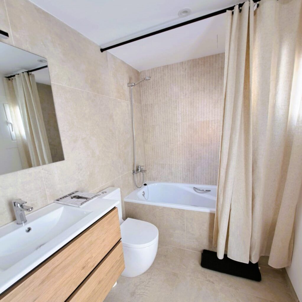 Moderno cuarto de baño con azulejos de color beige, una bañera con cabezal de ducha, cortina de ducha de color crema, tocador de madera con lavabo y espejo, y un inodoro. La luz natural procede de una pequeña ventana.