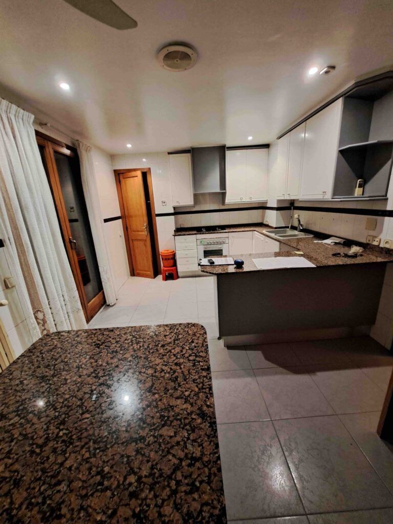 Cocina moderna con encimeras de granito, armarios blancos, suelo de baldosas y electrodomésticos empotrados. Una cortina cubre una puerta de cristal y al fondo se ve una puerta de madera. La cocina está bien iluminada con lámparas de techo.