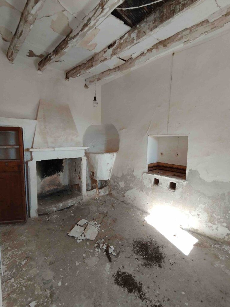 Una vieja cocina abandonada con el techo dañado, vigas de madera a la vista, una chimenea, un horno empotrado, paredes desconchadas y escombros esparcidos por el suelo agrietado. La luz del sol entra por una ventana e ilumina parte de la habitación.