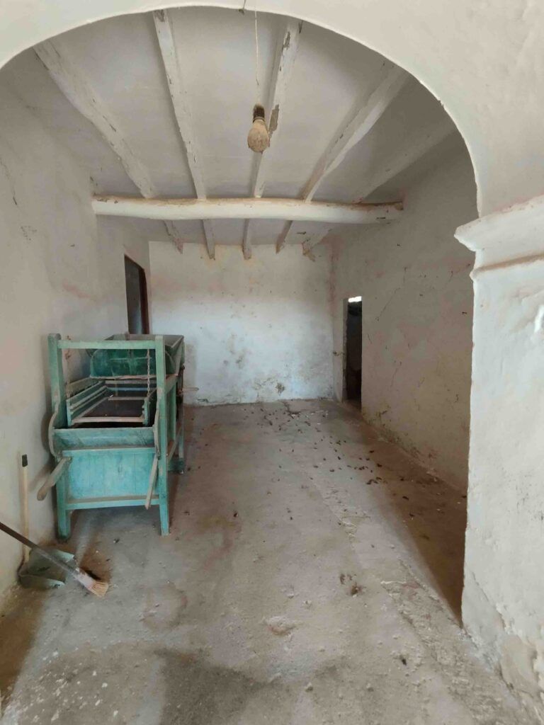 Una habitación polvorienta y vacía con paredes blancas, techo arqueado, vigas de madera a la vista y una sola bombilla. A la izquierda hay una vieja máquina de color turquesa y una escoba apoyada en la pared. El suelo está sucio y lleno de basura.
