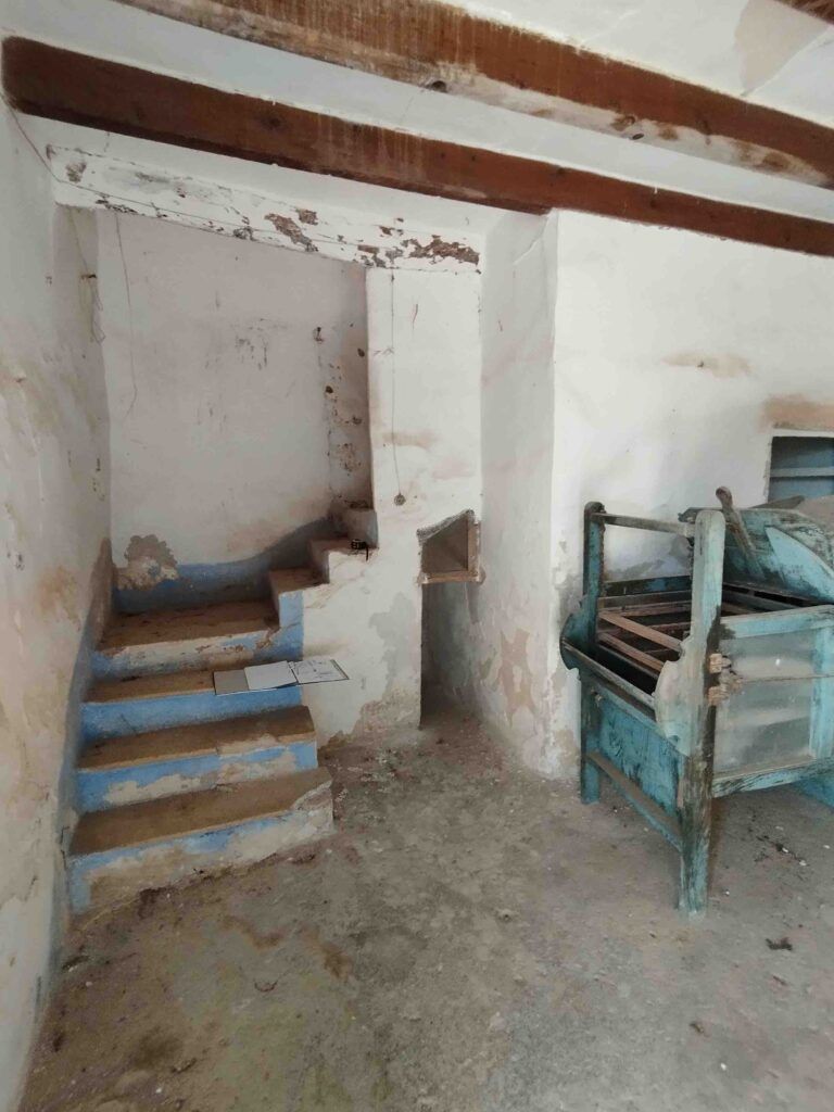 Unas viejas y desgastadas escaleras de hormigón con la pintura desconchada suben junto a un pequeño cubículo en una habitación destartalada. A la derecha hay una vieja máquina de madera azul. Las vigas del techo y las paredes agrietadas muestran signos de deterioro.
