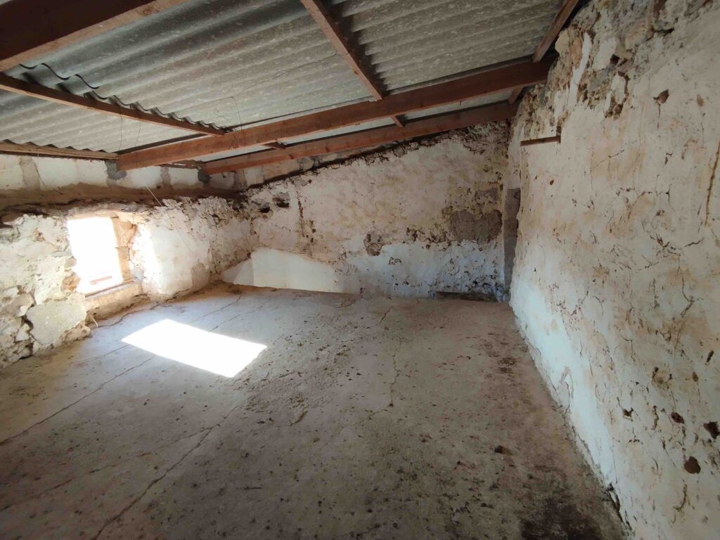 Una habitación vacía, rústica y abuhardillada, con toscas paredes de piedra encalada, una pequeña ventana por la que entra la luz del sol y un tejado de chapa ondulada sostenido por vigas de madera. El suelo parece polvoriento e inacabado.