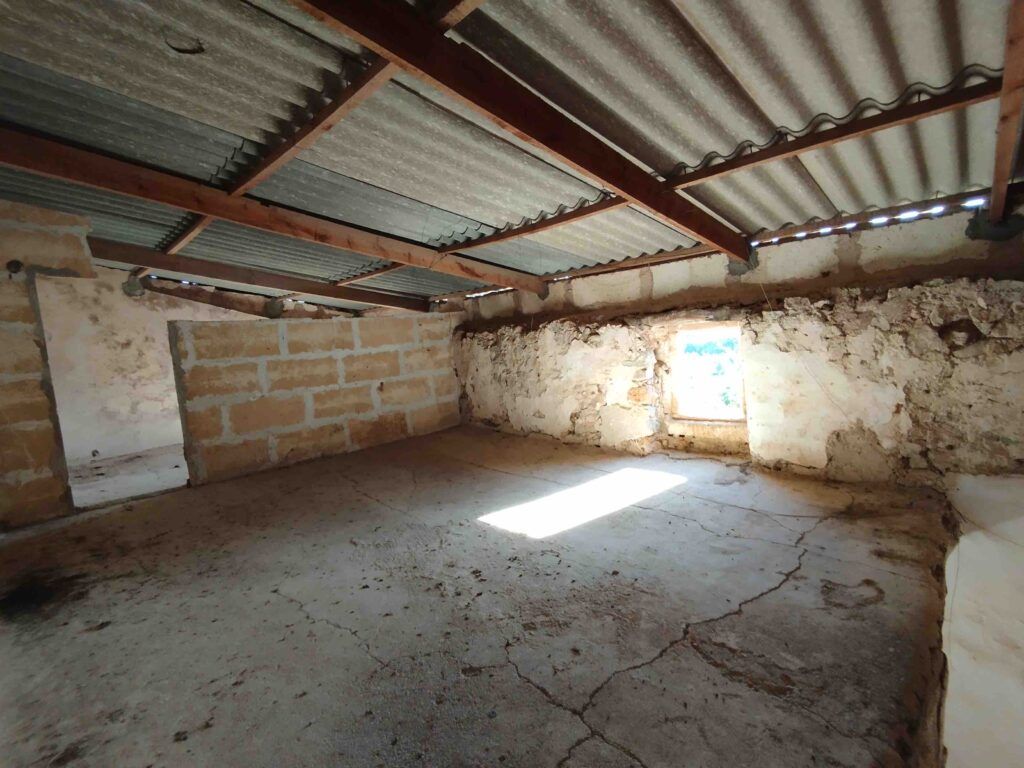 Una habitación vacía y rústica con suelos de hormigón agrietados, paredes de piedra tosca, una única ventana por la que entraba la luz del sol y un tejado de chapa ondulada sostenido por vigas de madera.