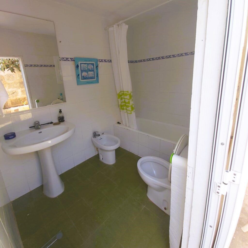Pequeño cuarto de baño con suelo de baldosas verdes, lavabo blanco, bidé, inodoro y bañera con cortina. En la pared de azulejos blancos, encima del lavabo, hay un espejo y un cuadro. La luz natural entra por una puerta abierta.
