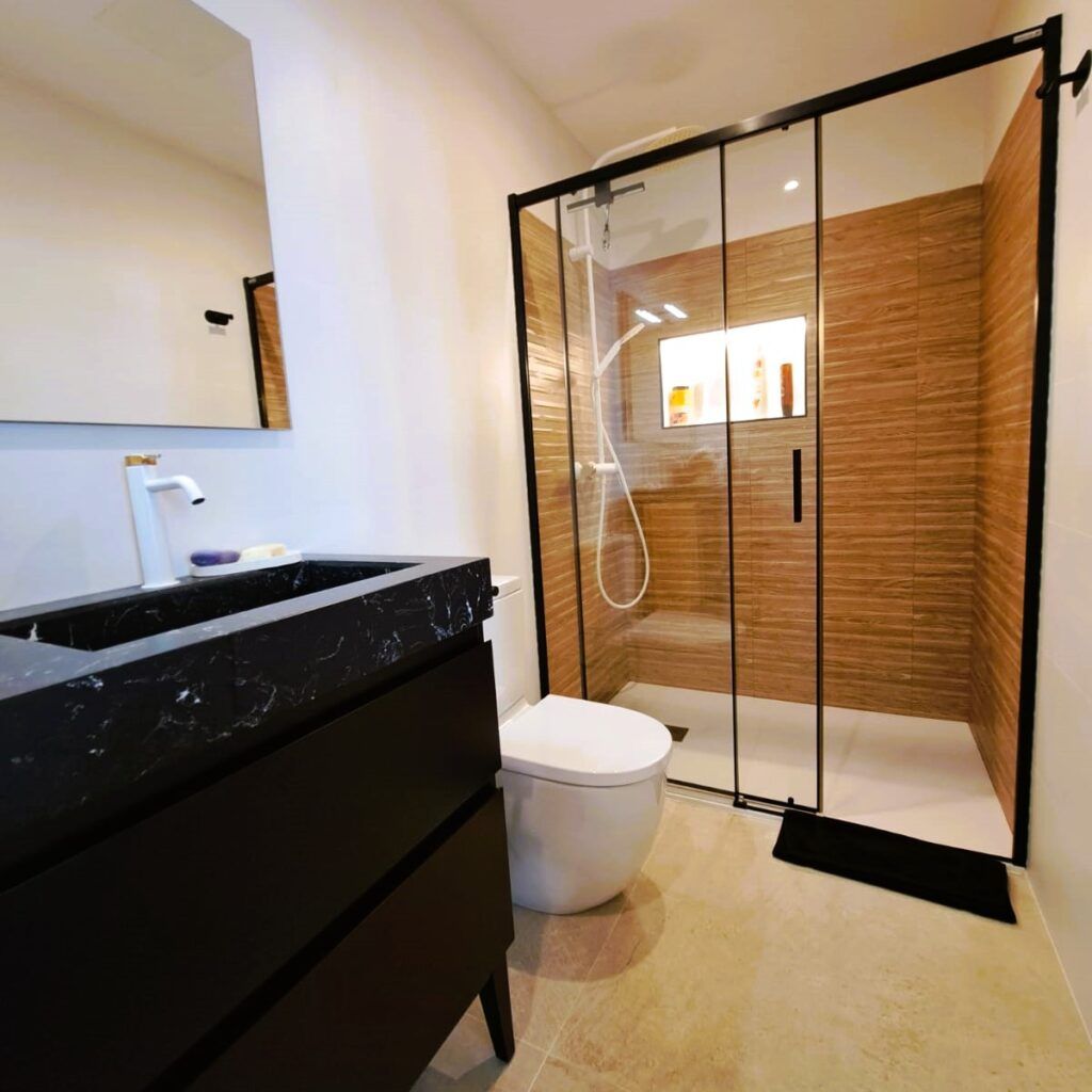 Moderno cuarto de baño con un tocador negro, un lavabo blanco, un inodoro montado en la pared y una ducha acristalada con azulejos marrones texturizados y una repisa empotrada. El suelo es de baldosas beige y el espacio es luminoso y limpio.