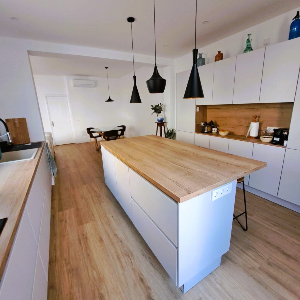 Cocina moderna con suelos de madera clara, una gran isla de madera, armarios blancos, lámparas colgantes negras y electrodomésticos empotrados. Al fondo se ve la zona de comedor con una pequeña mesa y sillas.