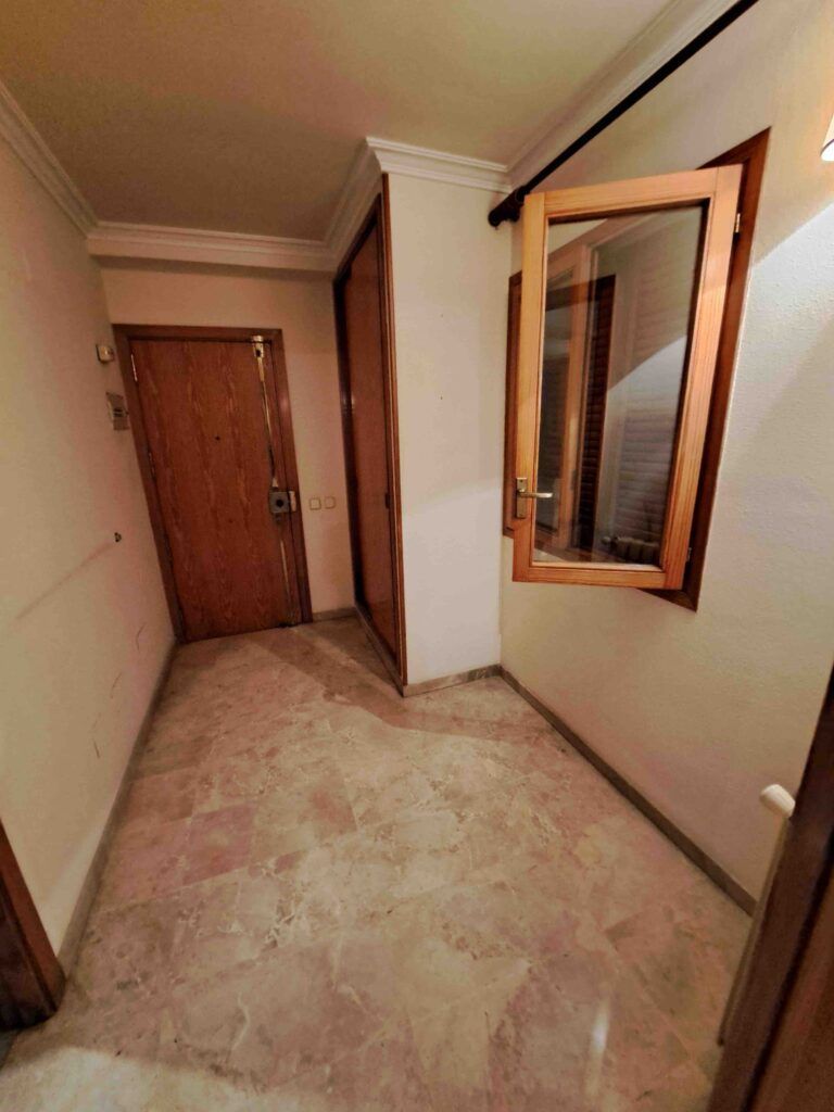 Pasillo con suelo de mármol, puertas de madera a la izquierda y al fondo, y una ventana abierta con marco de madera en la pared derecha. Las paredes son de color crema y la iluminación es cálida.