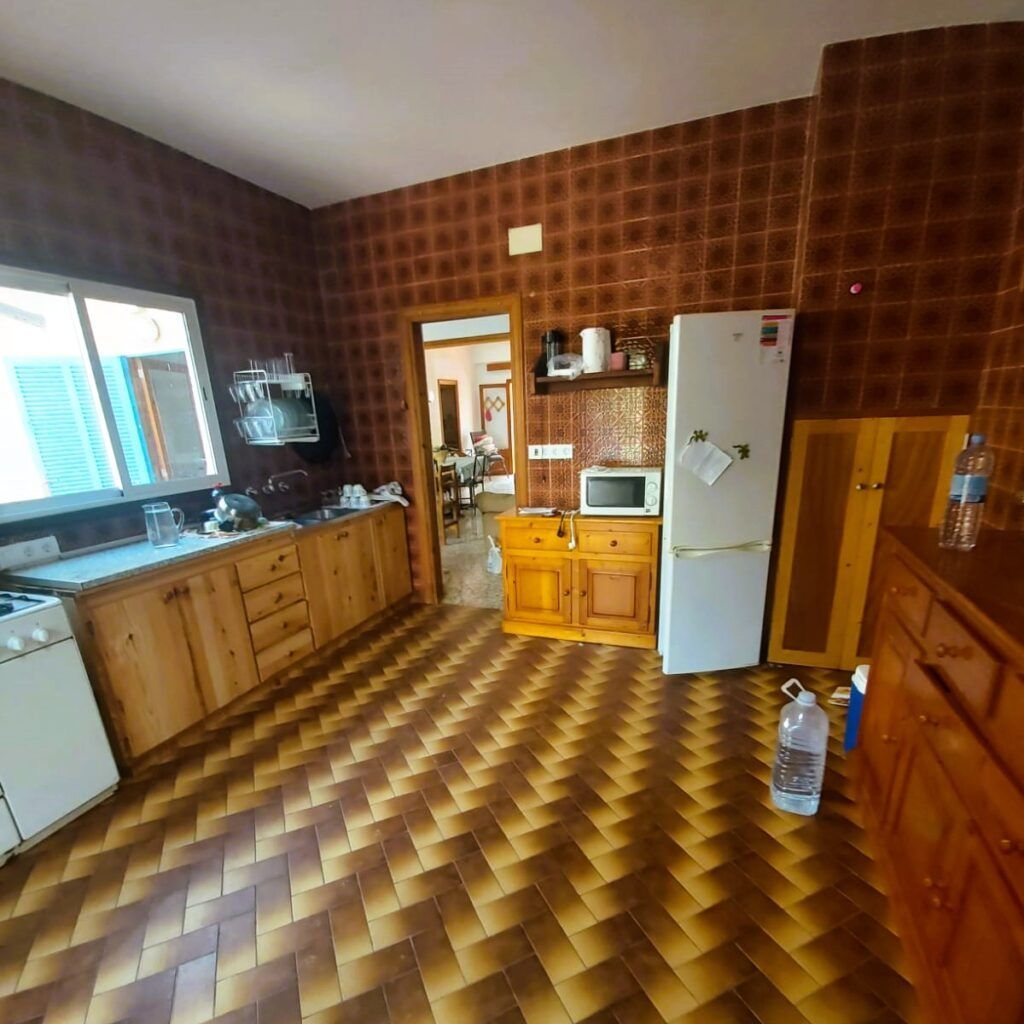 Cocina con paredes y suelo de baldosas marrones, armarios de madera, frigorífico blanco, microondas, fregadero, escurreplatos y una gran ventana. Una puerta da a un comedor; hay botellas de agua en el suelo y en la encimera.