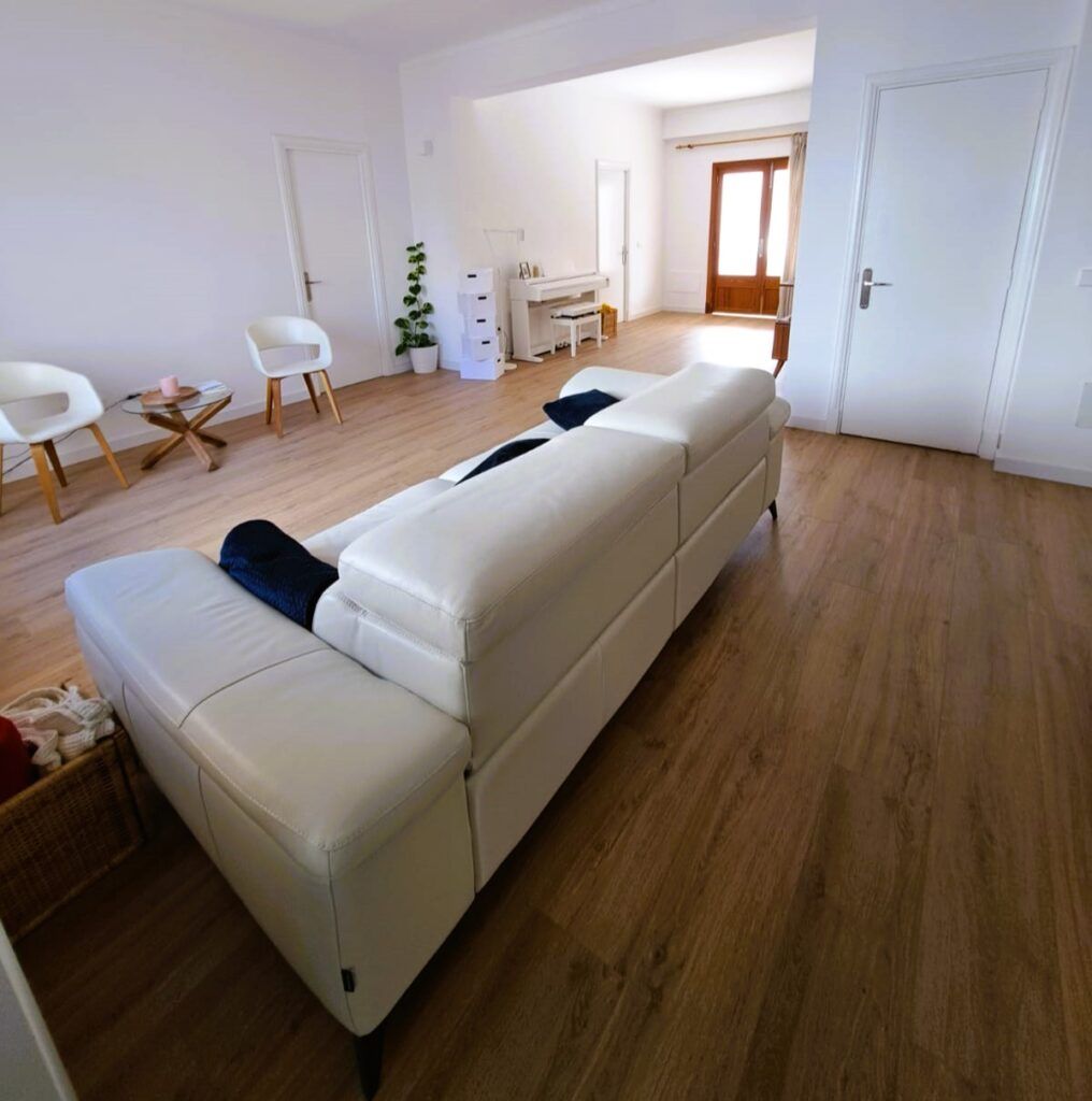Un salón luminoso y minimalista con un sofá blanco, dos sillas blancas, una pequeña mesa redonda, suelos de madera clara, paredes blancas y una puerta que da a otra zona por la que entra luz natural.