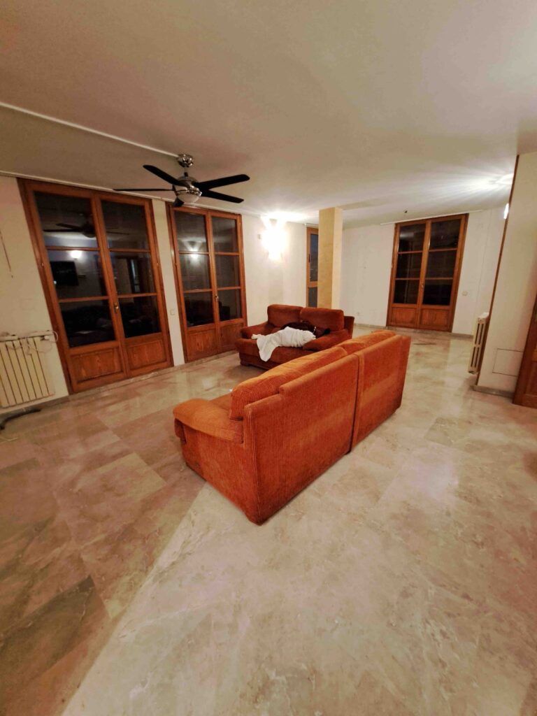 Un amplio salón con suelo de mármol, grandes ventanas con marcos de madera, dos sofás naranjas dispuestos en forma de L, un ventilador de techo y una suave iluminación cenital. Una manta blanca cubre uno de los sofás.