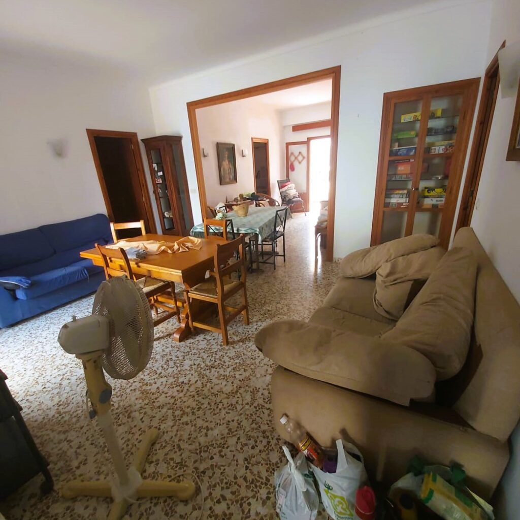 Una sala de estar con un sofá azul, un sofá beige, una mesa de comedor de madera con sillas, un ventilador de pie y una vitrina llena de juegos de mesa. La luz del sol entra por una habitación del fondo. El suelo es de baldosas.