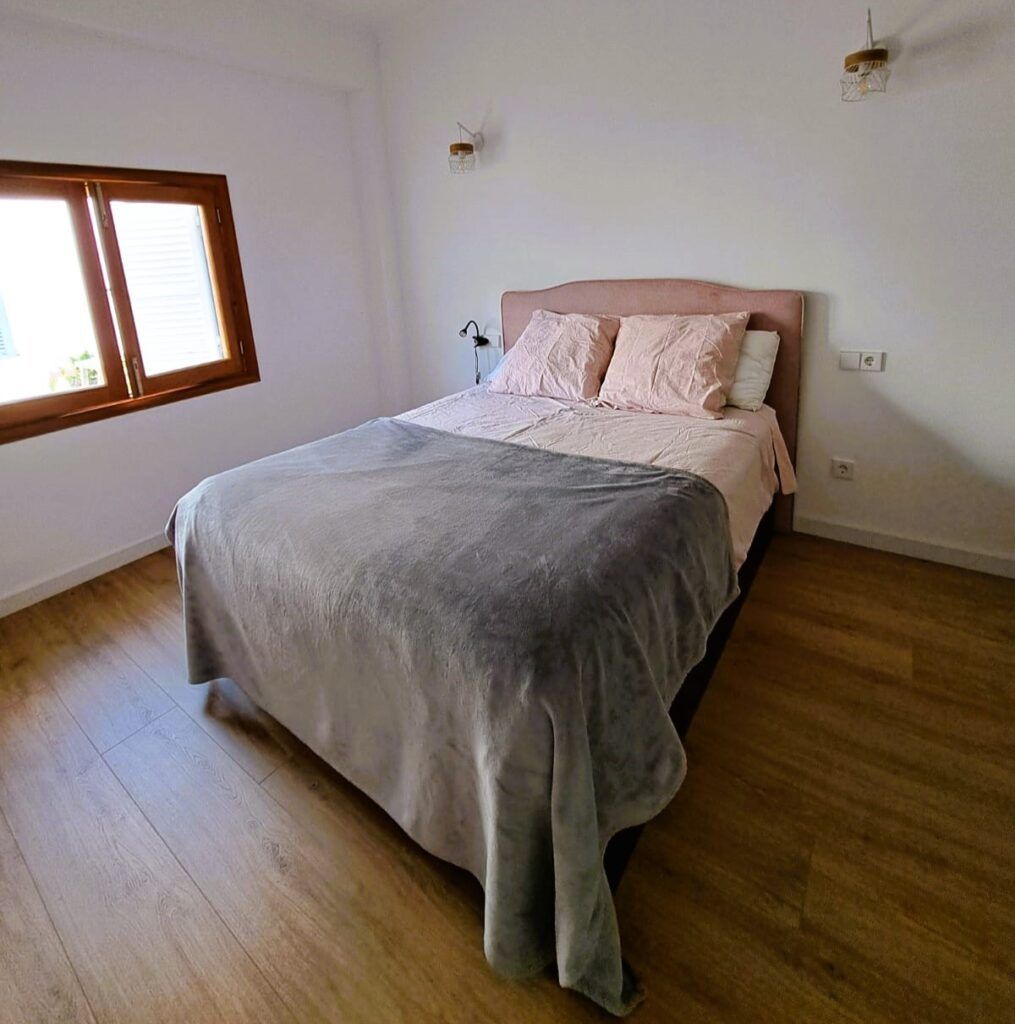 Una habitación ordenada con una cama doble cubierta con ropa de cama gris y rosa, colocada contra una pared blanca. Suelo de madera, ventana a la izquierda que deja pasar la luz y decoración minimalista.