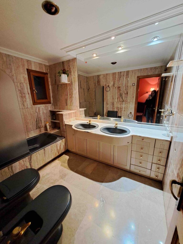 Un amplio cuarto de baño con paredes y suelo de mármol, con una bañera negra, un lavabo doble, un inodoro y un bidé negros, y un gran espejo. En la puerta se ve a una persona reflejada en el espejo.