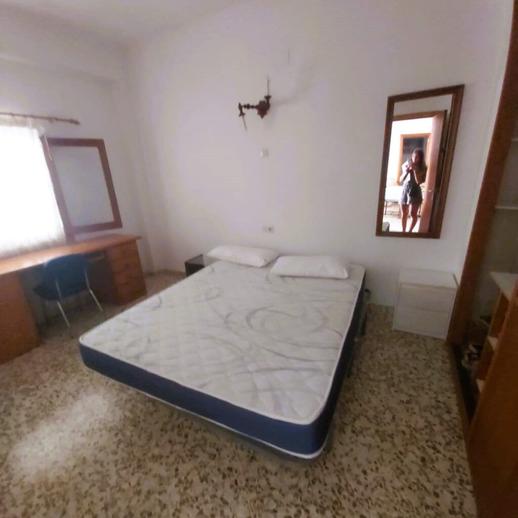 Un dormitorio con un colchón desnudo, dos almohadas, un escritorio bajo una ventana con cortinas, dos espejos y suelo de baldosas. Se ve a una persona haciendo la foto en el reflejo del espejo.