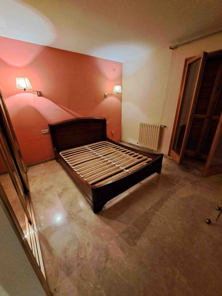 Dormitorio con somier de madera sin colchón, suelo de mármol, pared de acento rosa con dos lámparas de pared, radiador, armario con espejo y puertas de persiana de madera parcialmente abiertas.