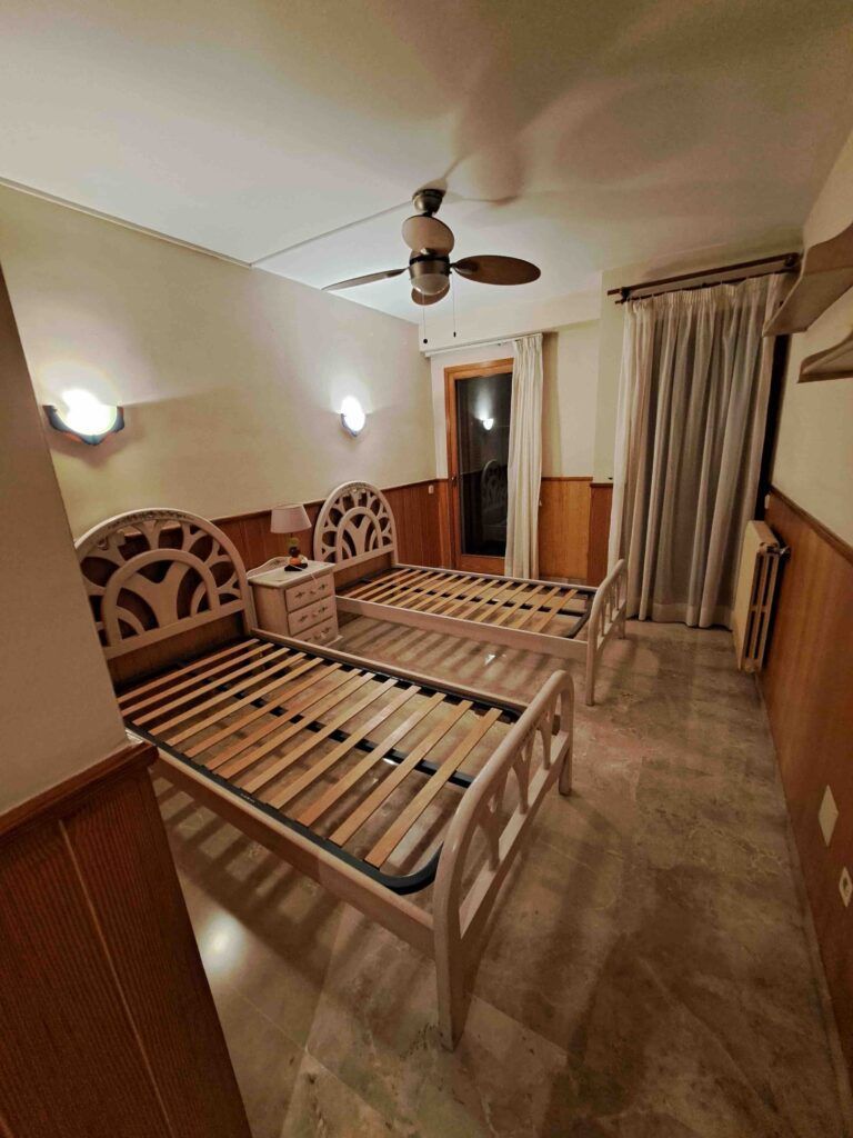 Dos camas individuales vacías con cabeceros decorativos están colocadas en paralelo en un dormitorio con suelo de mármol, mesilla de noche entre ellas, lámparas de pared, ventilador de techo, cortinas y una puerta que da a un balcón.