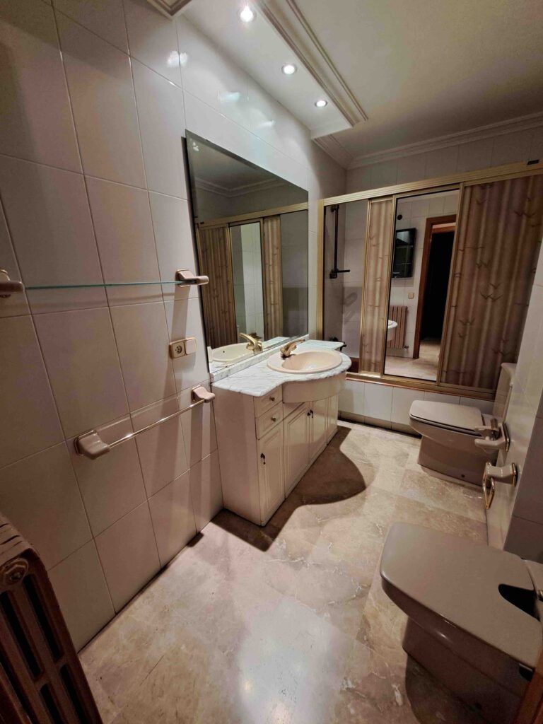 Un cuarto de baño con paredes alicatadas en blanco, un gran espejo sobre el lavabo y la encimera, baldosas beige en el suelo, un inodoro y un bidé, toalleros y armarios bajo el lavabo. Las luces del techo iluminan el espacio.