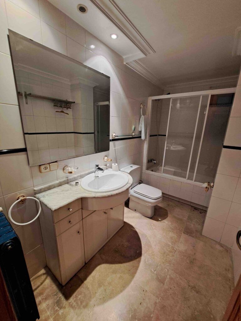 Un cuarto de baño limpio y alicatado, con un gran espejo, un lavabo blanco con armarios, un inodoro, un bidé y una ducha acristalada. Junto al lavabo cuelga una toalla, y el suelo parece ser de mármol beige.