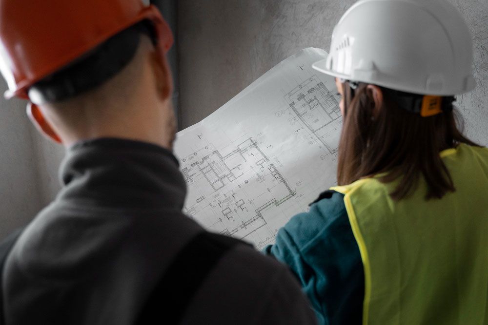 Dos personas con cascos y chalecos de seguridad examinan un gran plano arquitectónico, discutiendo los planes de construcción en un entorno de construcción interior.