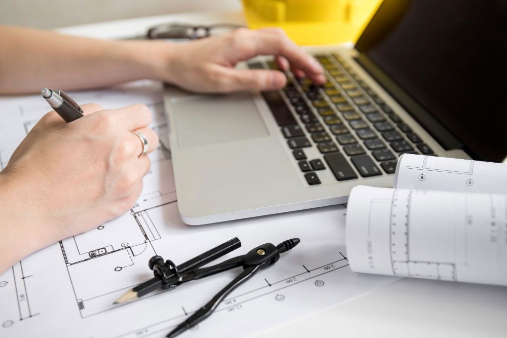 Persona trabajando en un escritorio con planos arquitectónicos, utilizando un ordenador portátil y escribiendo con un bolígrafo. Sobre el escritorio también se ven un plano enrollado, una brújula y un casco amarillo.