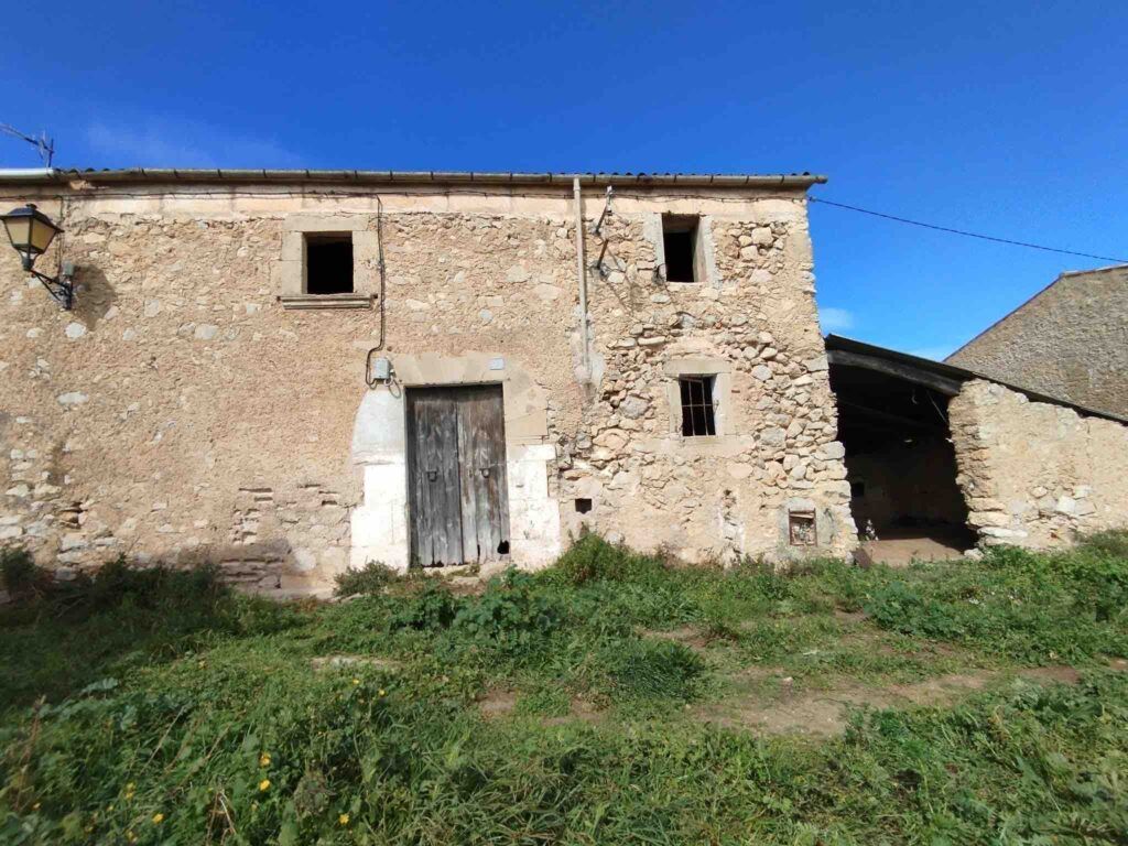 Antigua granja de piedra con puertas de madera y pequeñas ventanas cuadradas, rodeada de hierba crecida, bajo un cielo azul despejado. Una lámpara está montada en el lado izquierdo del edificio.