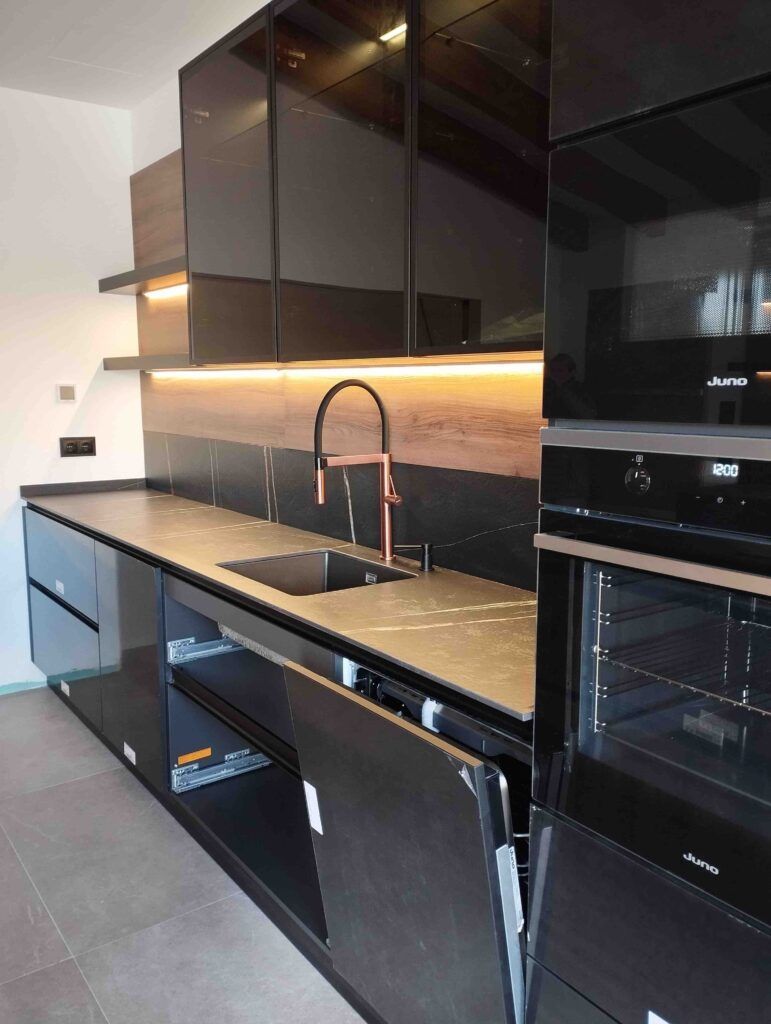 Cocina moderna con armarios negros, horno empotrado y fregadero con grifo alto negro. Algunas puertas de armarios y cajones están abiertos, mostrando un lavavajillas y espacios de almacenamiento. La iluminación bajo los armarios ilumina la encimera.