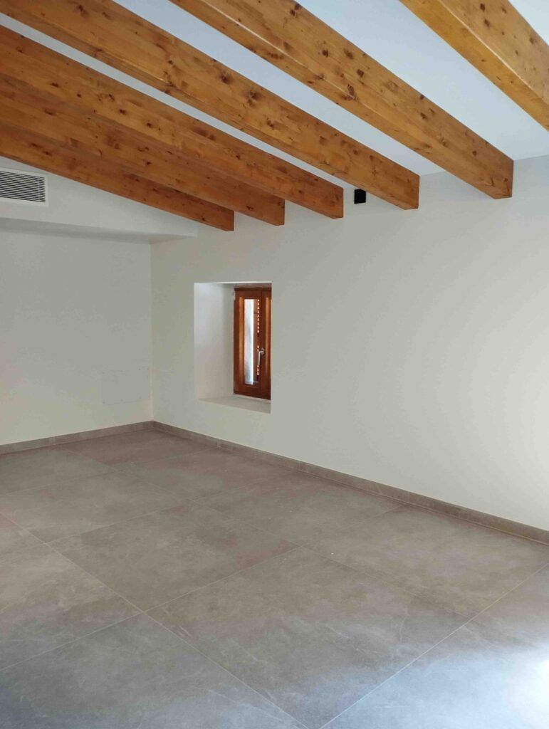 Una habitación luminosa y vacía con paredes claras, grandes baldosas grises en el suelo, vigas de madera vistas en el techo y una pequeña ventana con molduras de madera que deja pasar la luz natural.