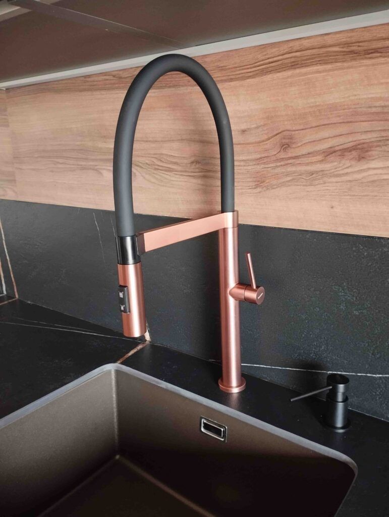 Junto al fregadero se instala una moderna grifería de cocina con acabado en negro mate y oro rosa, sobre una encimera negra y un protector contra salpicaduras con textura de madera.