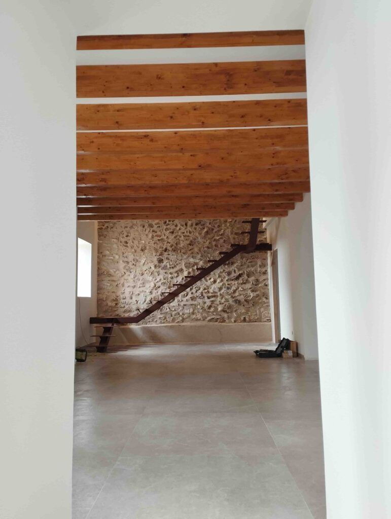 Interior espacioso con suelo de baldosas, paredes blancas, vigas de madera vistas en el techo y un muro de piedra al fondo con una sencilla escalera de madera. La luz natural entra por una ventana a la izquierda.