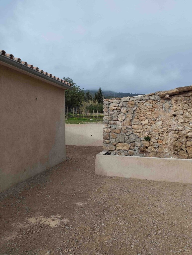 Un edificio beige y un muro de piedra se levantan sobre un camino de grava, con un bordillo bajo de hormigón. Al fondo, vegetación, árboles y un cielo nublado con nubes bajas.