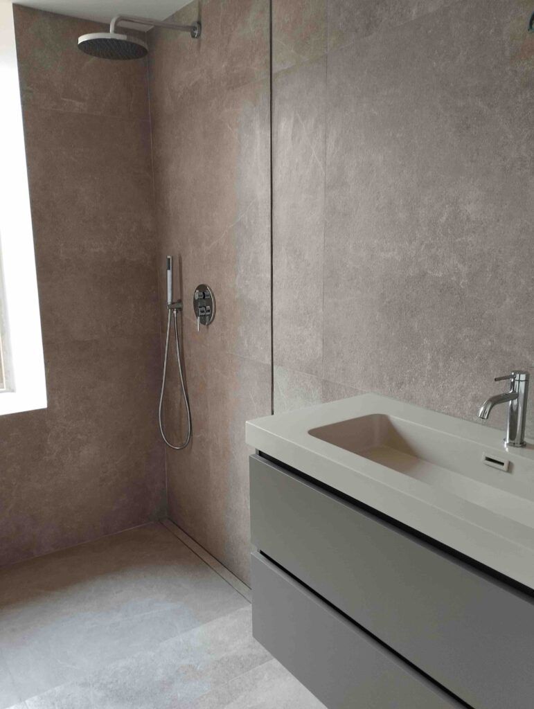 Cuarto de baño minimalista con grandes baldosas de color beige, una ducha a ras de suelo con cabezal de ducha tipo lluvia, ducha de mano y un lavabo moderno con pila rectangular y grifería cromada. El espacio es limpio y despejado.