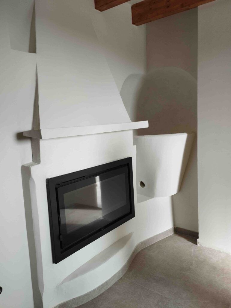Una moderna chimenea empotrada con una puerta de cristal con marco negro está encastrada en una pared lisa y esculpida en blanco. La zona circundante presenta líneas limpias y minimalistas y suelos de colores neutros.