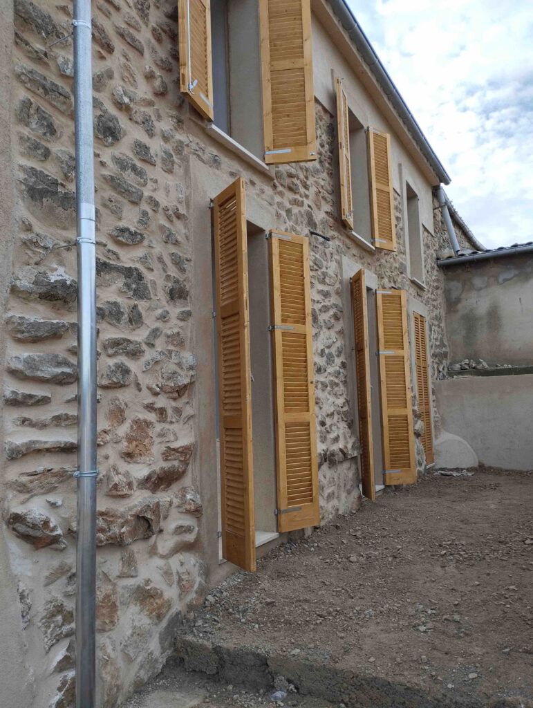 Una casa de piedra con cuatro juegos de contraventanas de madera abiertas en sus ventanas y puertas. El suelo es de tierra desnuda y un desagüe metálico recorre el lado izquierdo del edificio bajo un cielo parcialmente nublado.