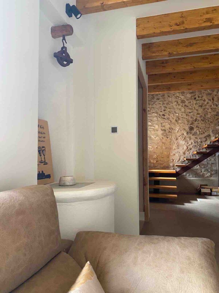 Una acogedora sala de estar con un sofá beige en primer plano, vigas de madera vistas en el techo, una pared de piedra y una escalera de madera que conduce al piso superior. Un pequeño objeto decorativo descansa sobre una repisa blanca junto a la pared.
