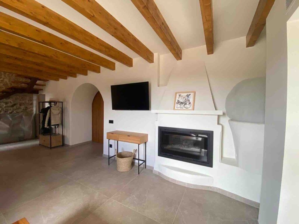 Moderno salón con vigas de madera vistas en el techo, un televisor montado en la pared, una chimenea, una mesita de madera con una cesta debajo y un perchero en la esquina. El suelo es de baldosas de color claro.