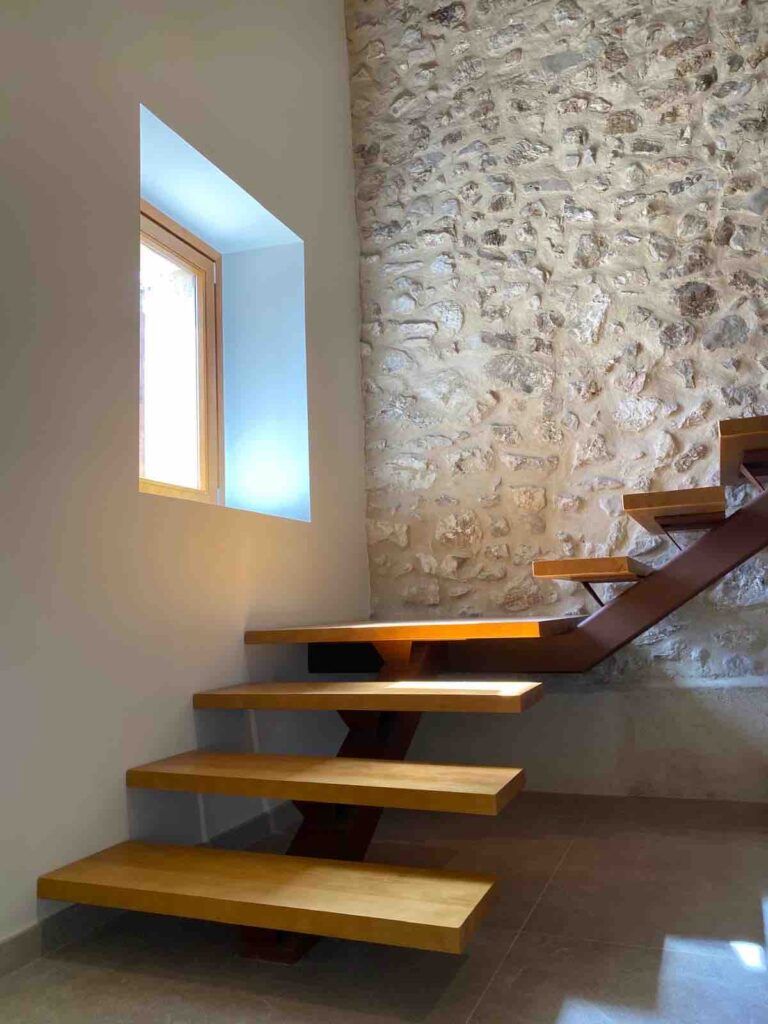 Una moderna escalera flotante de madera con contrahuellas abiertas se encuentra junto a una pared de piedra rústica y una ventana cuadrada que deja entrar la luz natural.