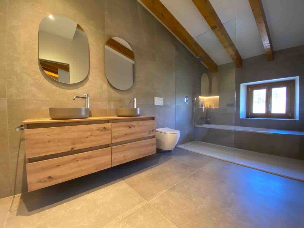 Moderno cuarto de baño con azulejos de color beige, dos espejos ovalados sobre un lavabo doble de madera, un inodoro montado en la pared, una ducha con mampara de cristal y una ventana que deja pasar la luz natural. Vigas de madera vistas en el techo.