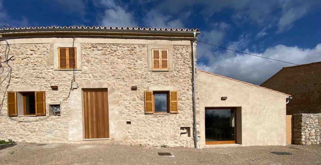 Una casa rústica de piedra con puertas y contraventanas de madera, enmarcada en un cielo parcialmente nublado. El suelo está sin pavimentar, y hay una pequeña extensión con una gran puerta de cristal en el lado derecho.