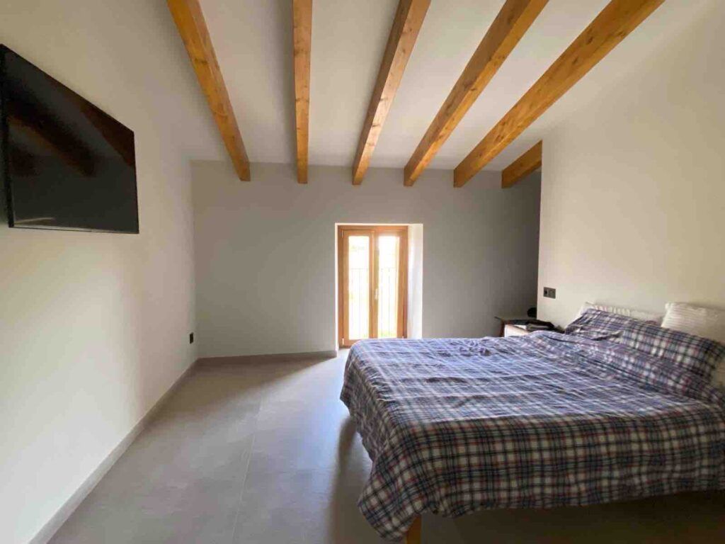 Un dormitorio luminoso con vigas de madera vistas en el techo, una cama doble cubierta con una colcha de cuadros escoceses, un televisor montado en la pared y una ventana que deja entrar la luz natural.