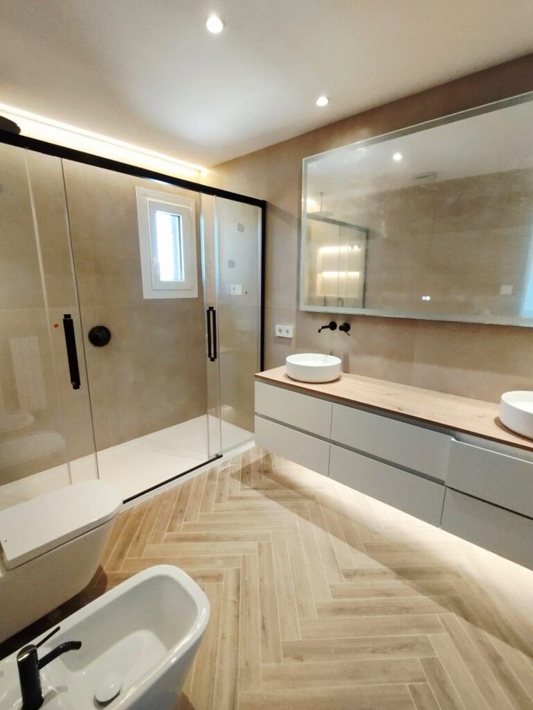 Moderno cuarto de baño con una gran ducha de cristal, dos lavabos redondos sobre una encimera de madera, un espejo de pared, un bidé y baldosas de madera clara en espiga. Las paredes son de color beige y la iluminación está empotrada.
