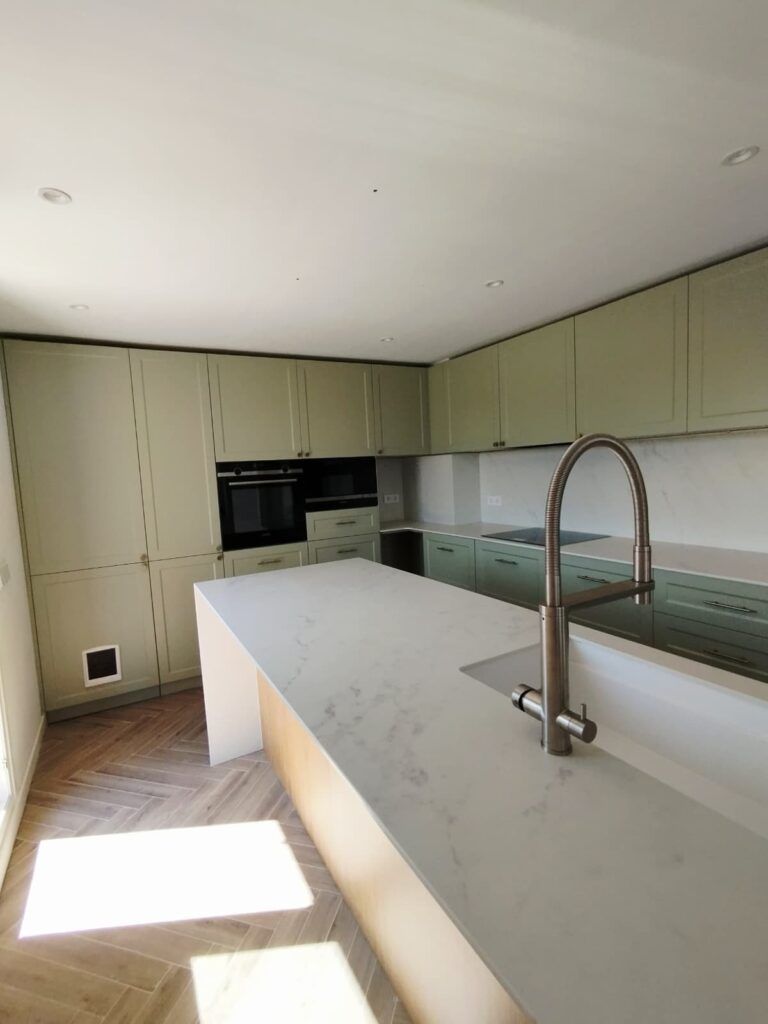 Cocina moderna con armarios de color verde claro, encimeras de mármol, horno negro empotrado, suelo de madera en espiga y una gran isla con un fregadero con grifo alto. La luz del sol entra a raudales por una ventana oculta.