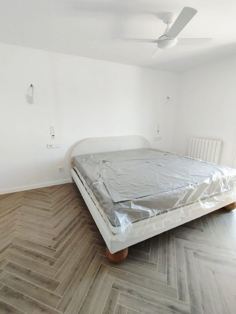 Un dormitorio minimalista con una cama grande cubierta de plástico, paredes blancas, suelo de madera con dibujo de espiga, un ventilador de techo, lámparas de pared y un radiador a la derecha.